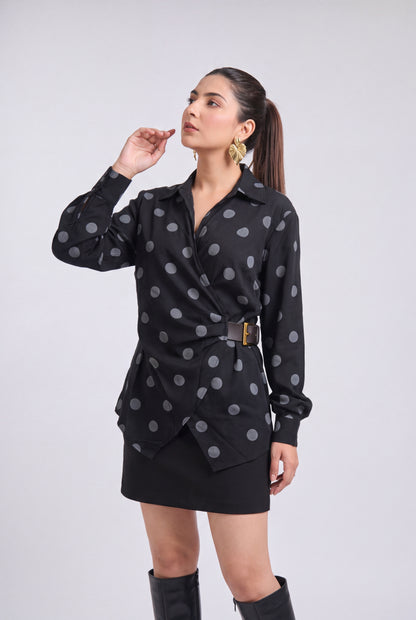 A woman modeling a black long-sleeve wrap-style blouse with gray polka dots and a matching black mini skirt, paired with black knee-high boots, against a light gray studio background.