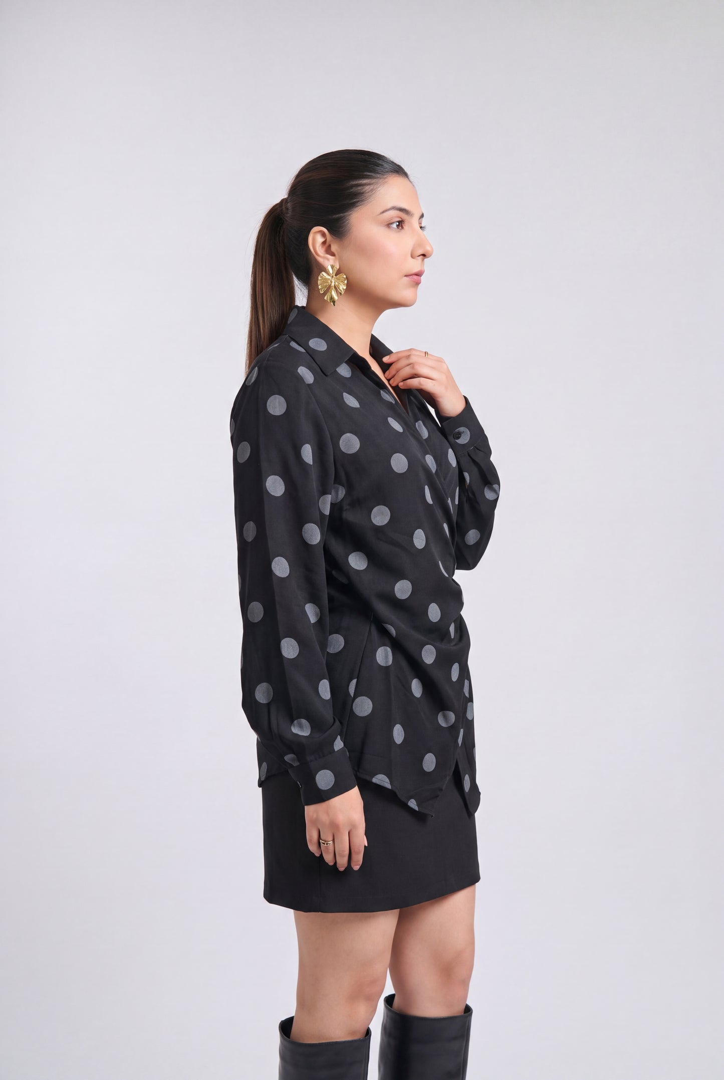 A woman modeling a black long-sleeve button-up shirt with a gray polka dot pattern, paired with a black mini skirt and black knee-high boots, set against a plain light gray studio background.