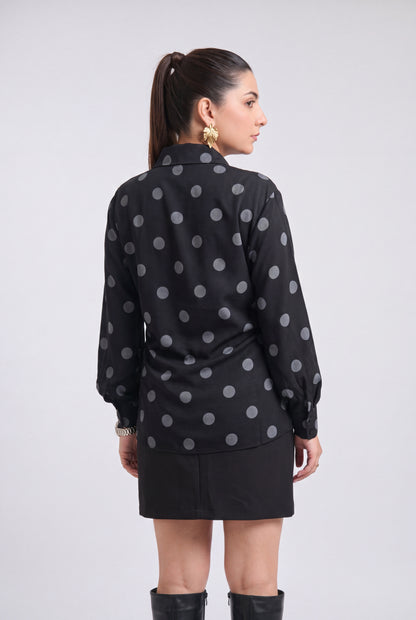 A woman shown from the back wearing a black long-sleeve blouse with a gray polka dot pattern and a black mini skirt, standing against a light gray studio background.