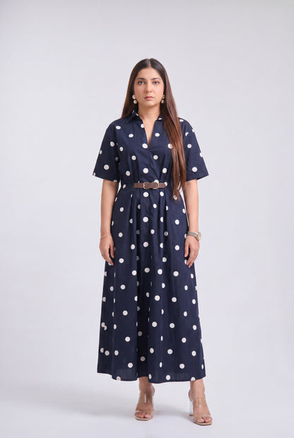 A woman modeling a navy blue short-sleeve maxi dress with white polka dots, featuring a matching brown belt, against a light gray studio background.
