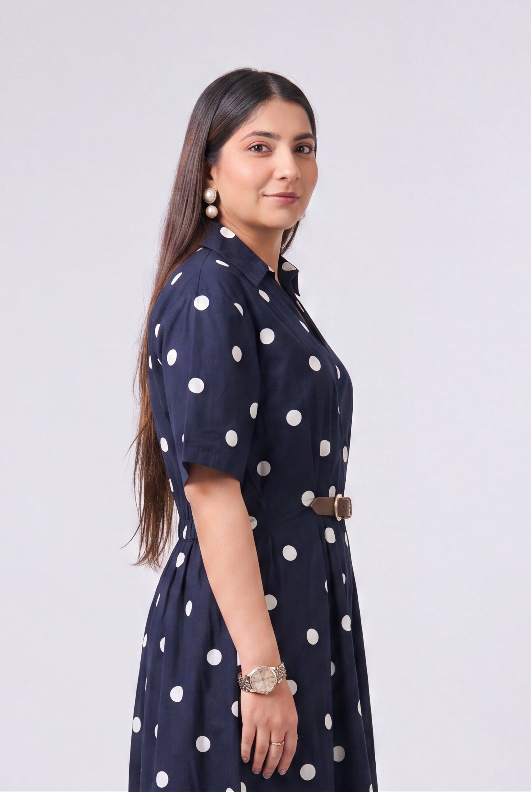 A woman modeling a navy blue midi dress with white polka dots, featuring short sleeves, a high collar, and a matching belt, set against a plain light gray studio background.
