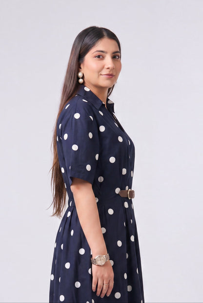 A woman modeling a navy blue midi dress with white polka dots, featuring short sleeves, a high collar, and a matching belt, set against a plain light gray studio background.