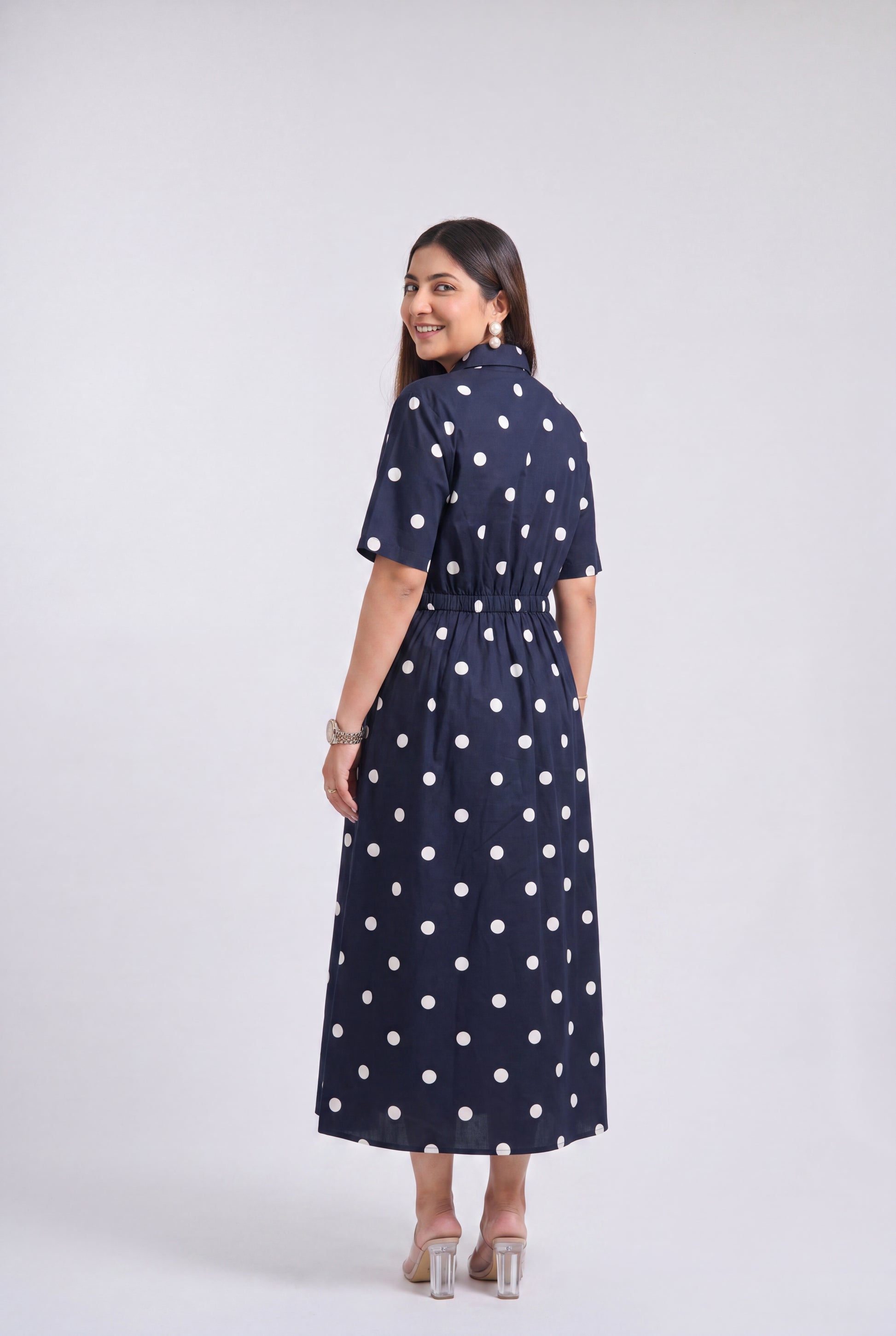 A woman modeling a navy blue midi dress with white polka dots, shown from the back against a light gray studio background.