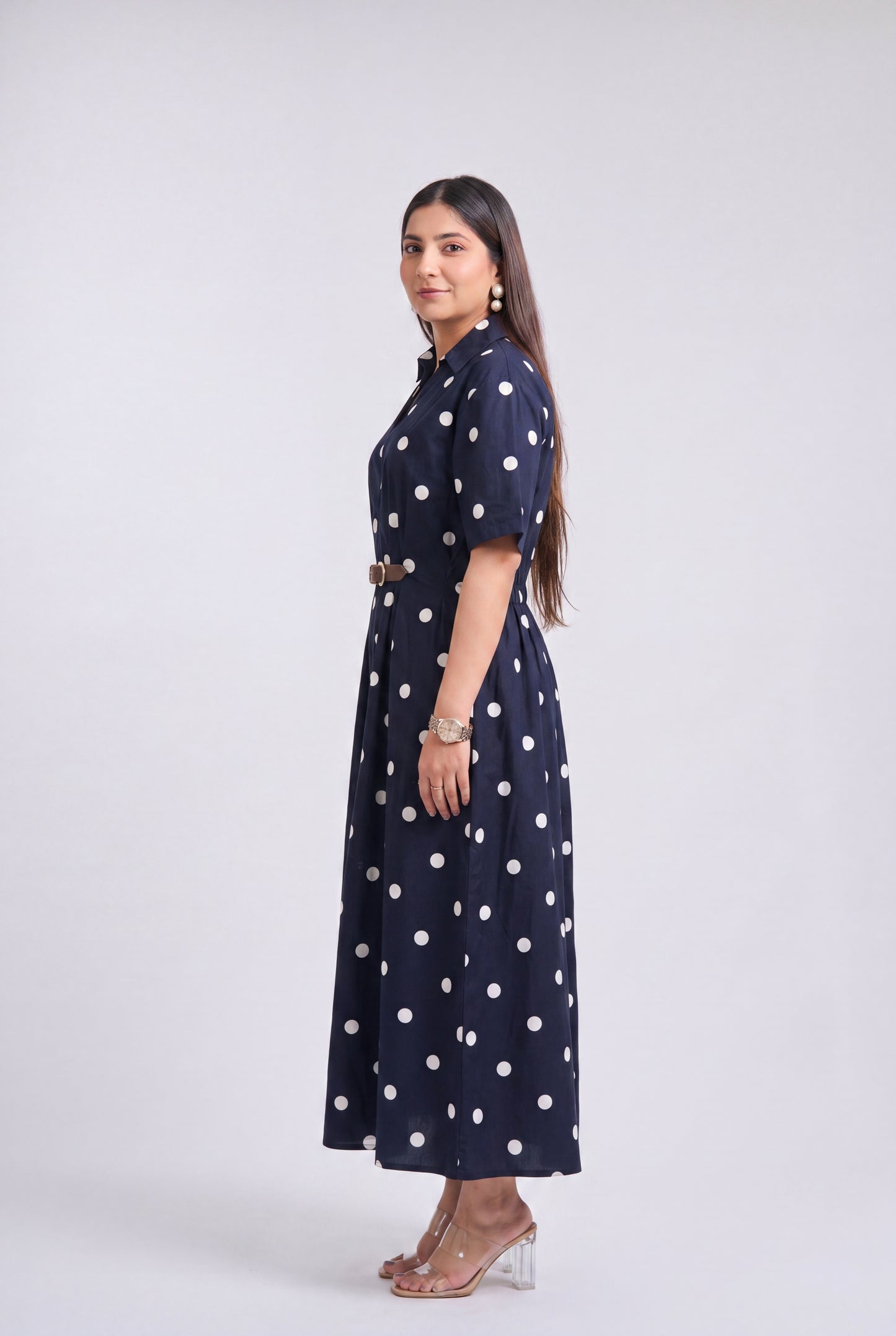 A woman modeling a navy blue midi dress with white polka dots, featuring short sleeves, a high collar, and a matching belt, set against a plain light gray studio background.