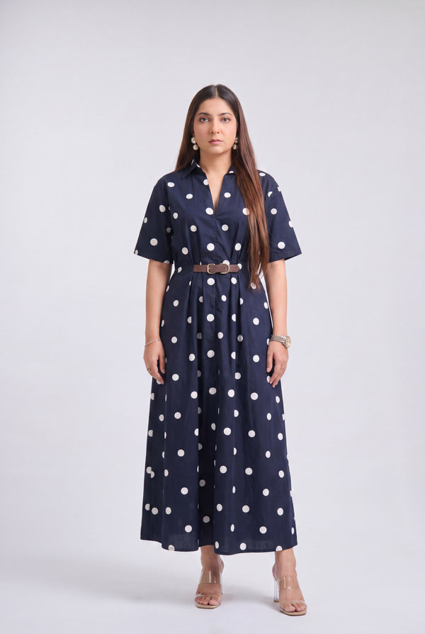 A woman modeling a navy blue short-sleeve maxi dress with white polka dots, featuring a matching brown belt, against a light gray studio background.