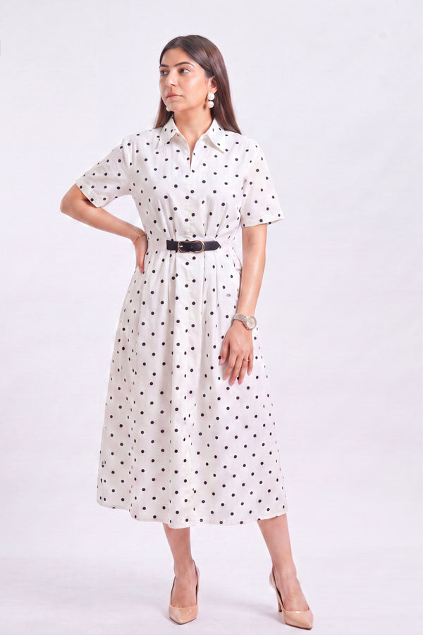 A woman modeling a white short-sleeve midi dress with black polka dots, featuring a pointed collar and a thin black belt at the waist, paired with beige heels against a light gray studio background.