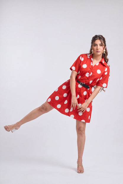 A woman modeling a red short-sleeve shirt dress with white polka dots and a black belt, posed dynamically against a light gray studio background.