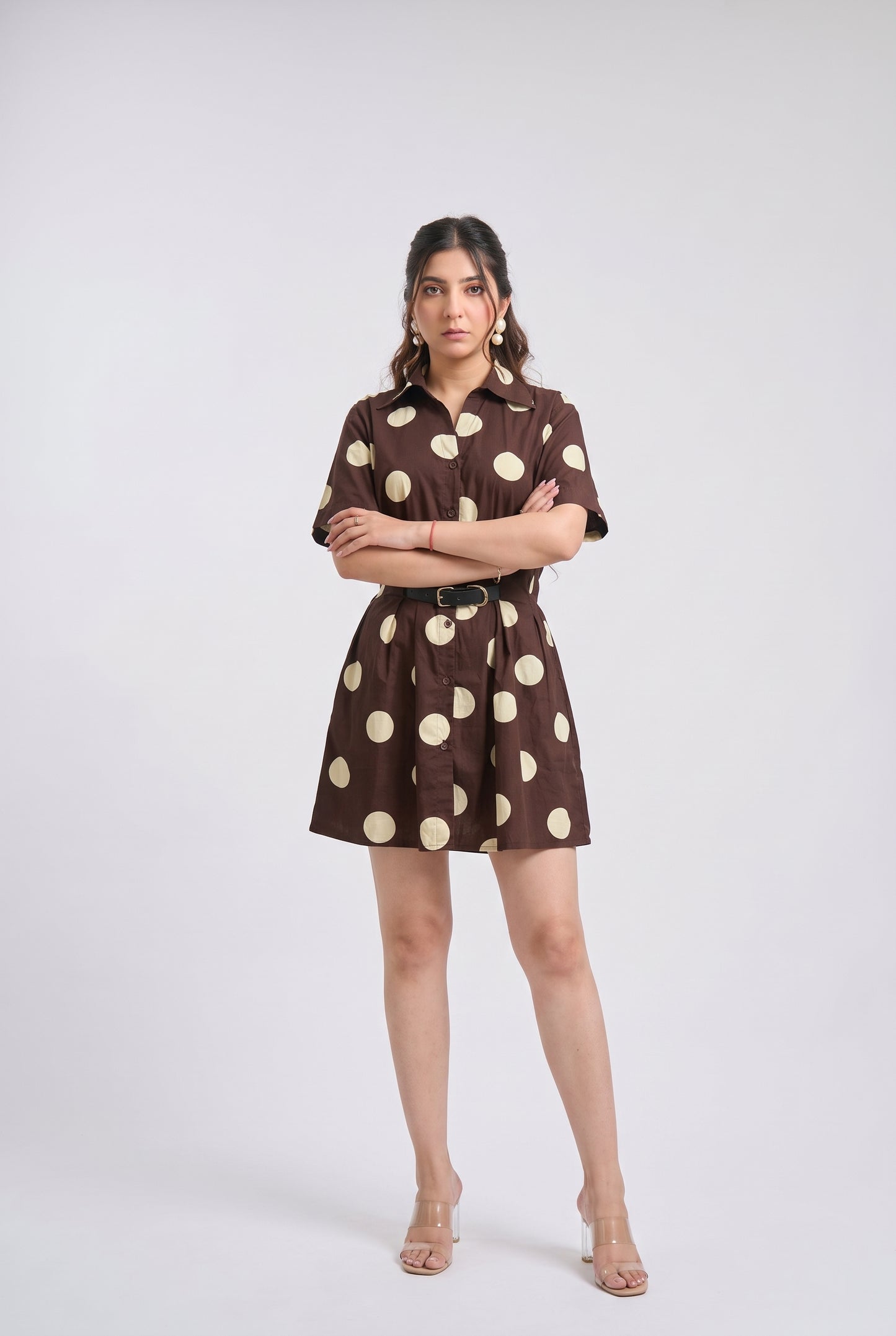 A woman modeling a brown short-sleeve mini dress with large beige polka dots, a pointed collar, and a matching belt, photographed against a light gray studio background.