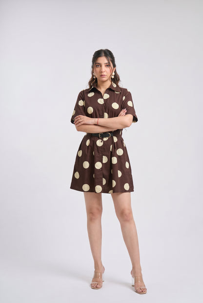 A woman modeling a brown short-sleeve mini dress with large beige polka dots, a pointed collar, and a matching belt, photographed against a light gray studio background.