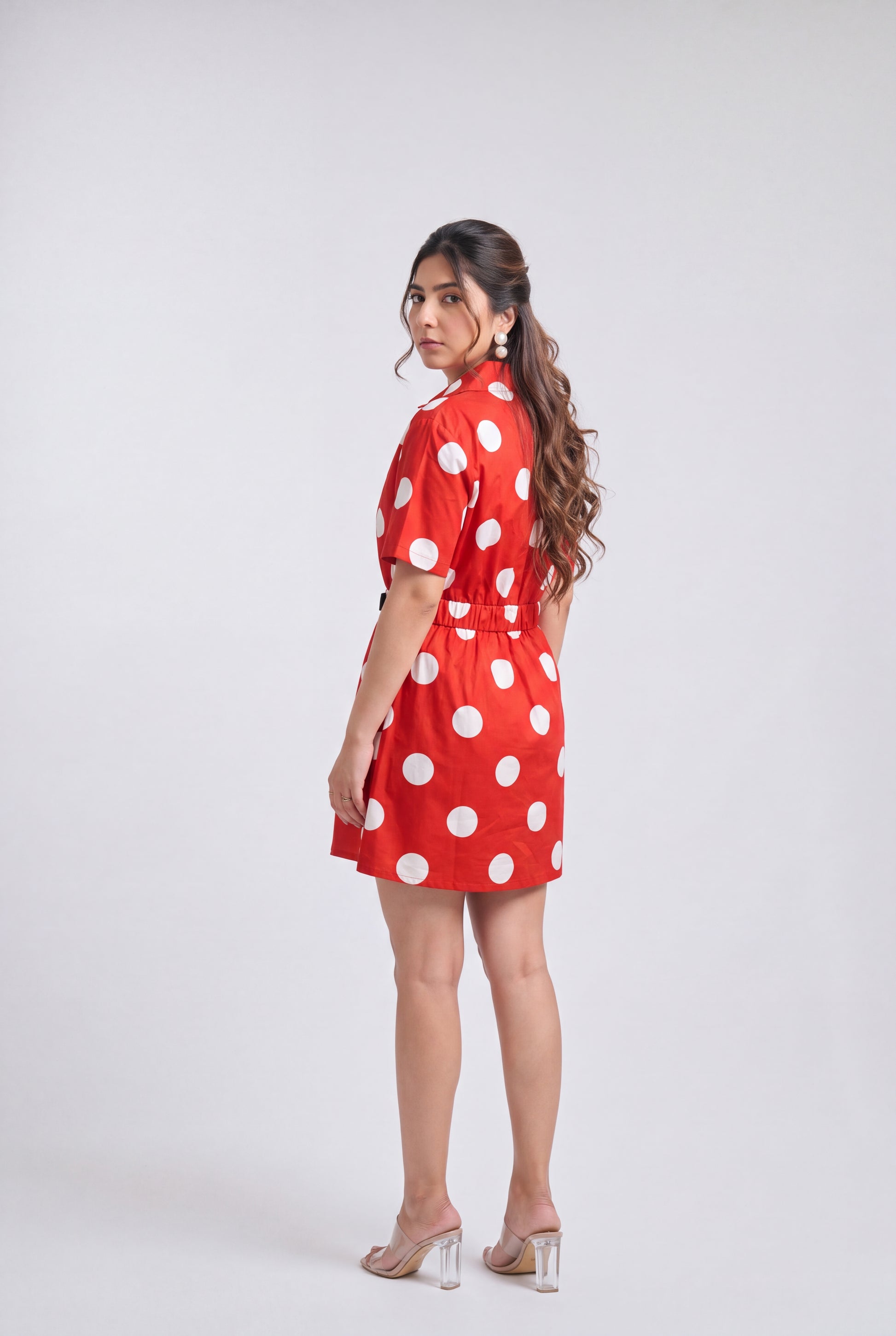 A woman modeling a red short-sleeve mini dress with large white polka dots, shown from a side-back angle against a light gray studio background.