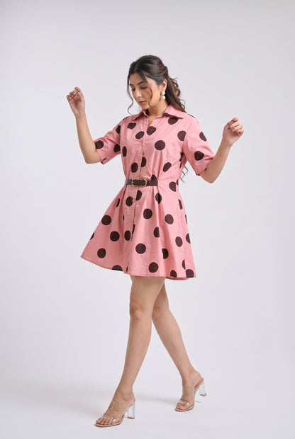 A woman modeling a pink short-sleeve mini dress with large black polka dots and a thin dark belt, posed against a light gray studio background.