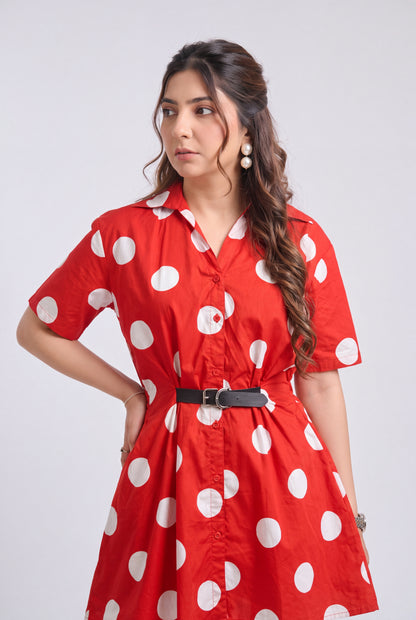 A woman modeling a red short-sleeve shirt dress with large white polka dots, featuring a pointed collar, button front, and a black belt at the waist, set against a plain light gray studio background.