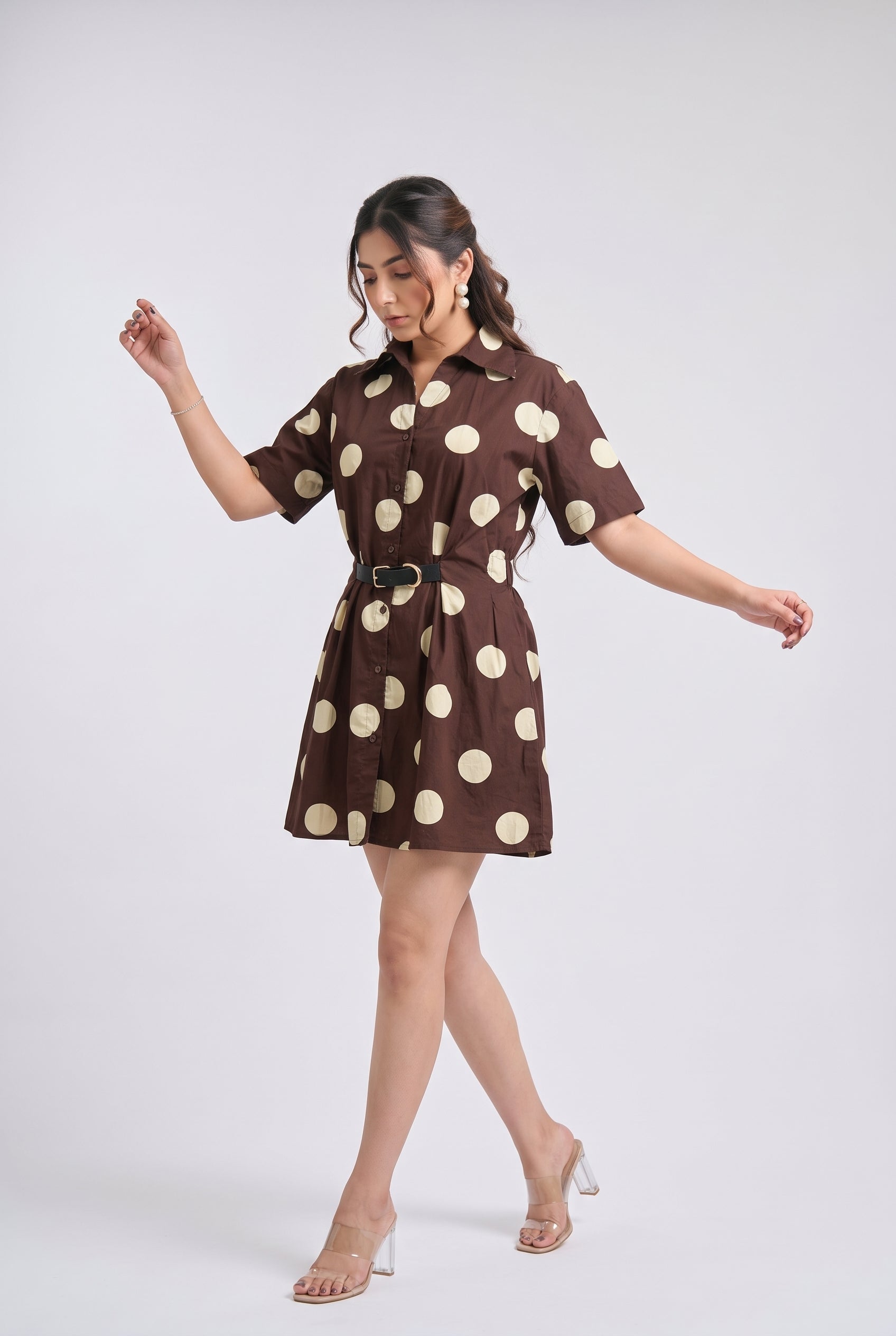 A woman modeling a brown short-sleeve mini dress with beige polka dots and a matching belt, posed against a light gray studio background.