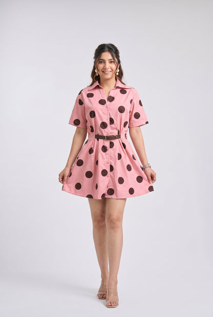 A woman modeling a pink short-sleeve collared dress with large dark brown polka dots and a thin dark belt, photographed against a light gray studio background.