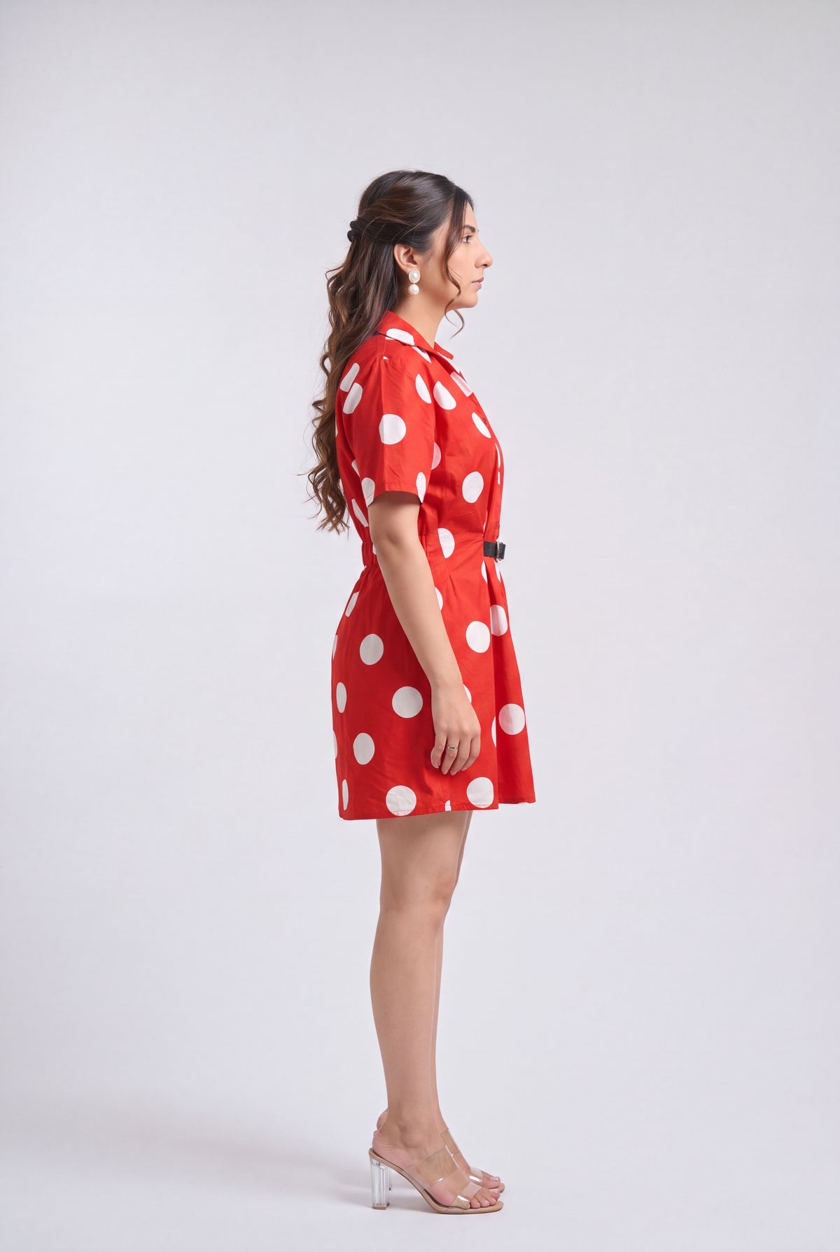 A woman in side profile wearing a red short-sleeve collared dress with large white polka dots and a matching belt, standing against a light gray studio background.