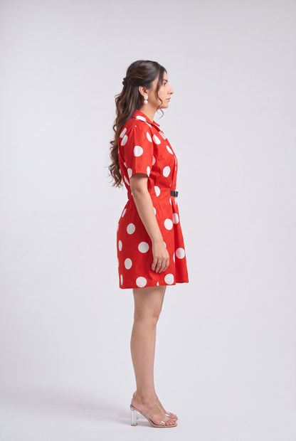 A woman in side profile wearing a red short-sleeve collared dress with large white polka dots and a matching belt, standing against a light gray studio background.