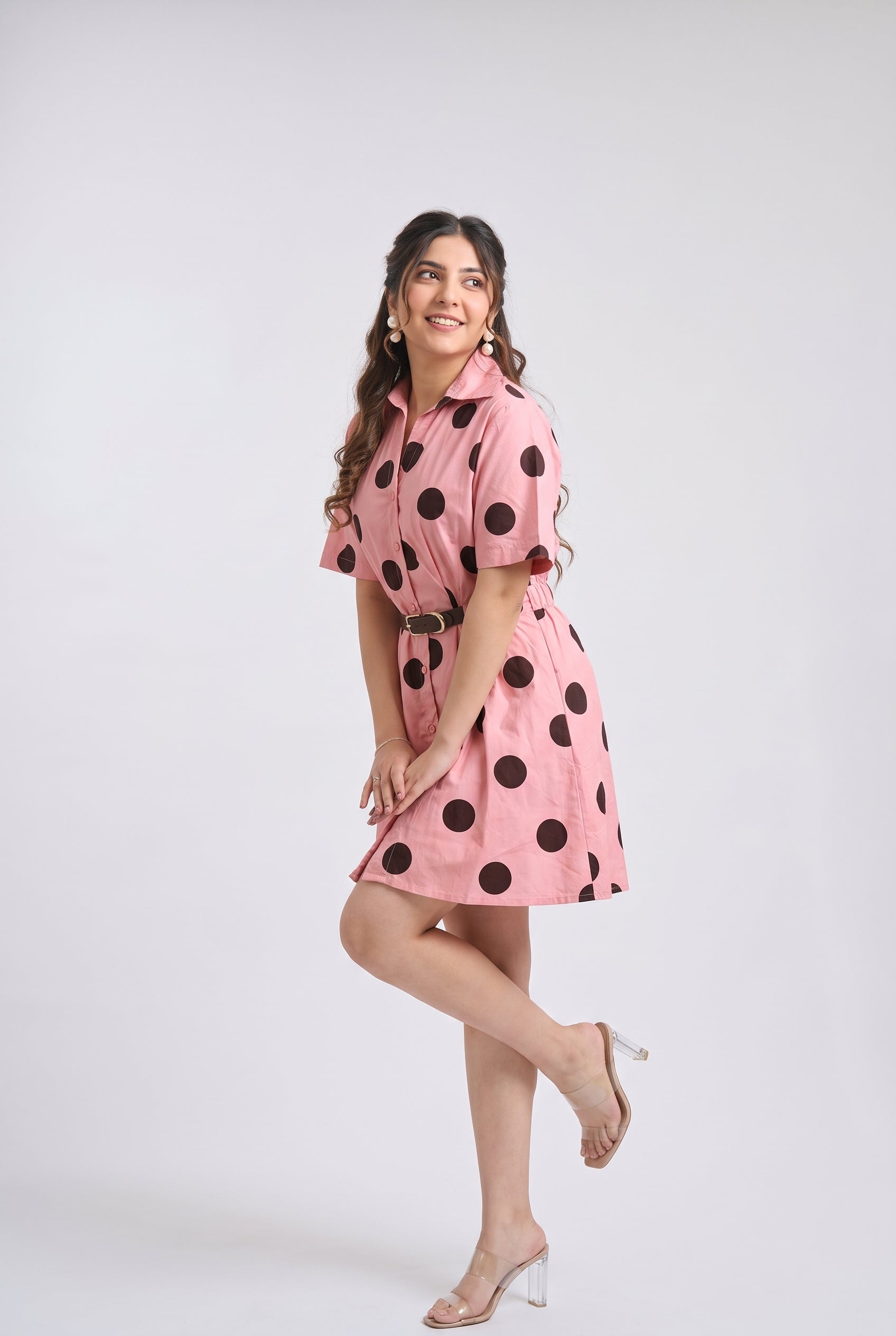 A woman modeling a pink short-sleeve collared dress with large black polka dots, standing against a light gray studio background.