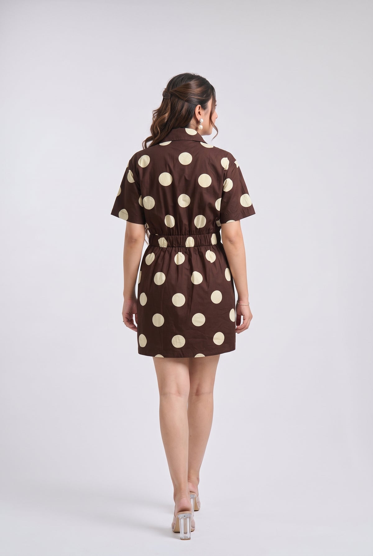 A woman viewed from behind wearing a brown short-sleeve mini dress with large cream polka dots and a tie waist, standing against a light gray studio background.