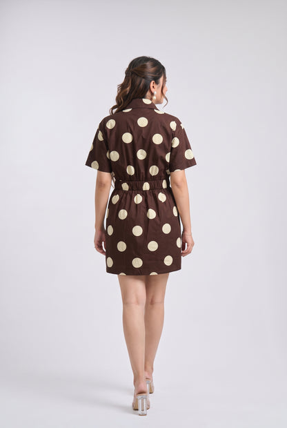 A woman viewed from behind wearing a brown short-sleeve mini dress with large cream polka dots and a tie waist, standing against a light gray studio background.