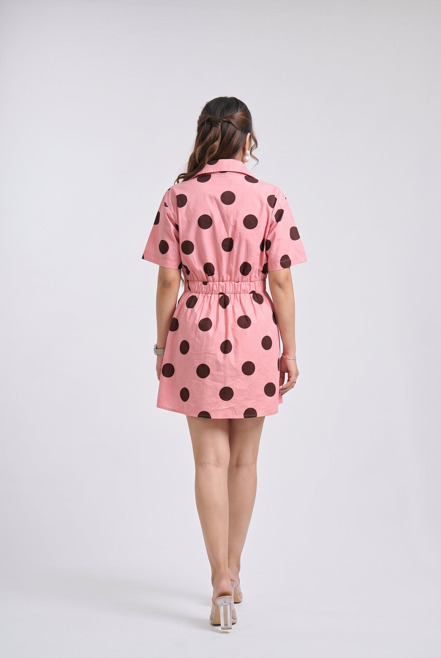A woman viewed from behind wearing a pink short-sleeve mini dress with large dark brown polka dots, standing against a light gray studio background.
