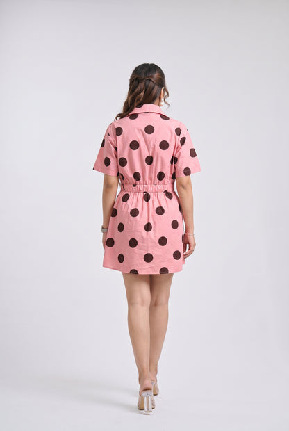 A woman viewed from behind wearing a pink short-sleeve mini dress with large dark brown polka dots, standing against a light gray studio background.