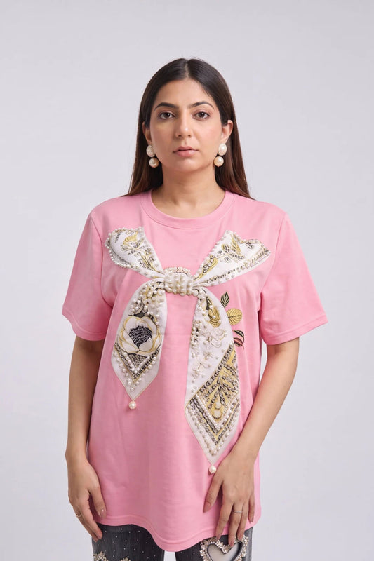 A woman modeling a pink t-shirt with a large decorative bow printed on the front, featuring a patterned scarf design in beige, white, and gold. She is wearing pearl earrings against a plain light gray background.