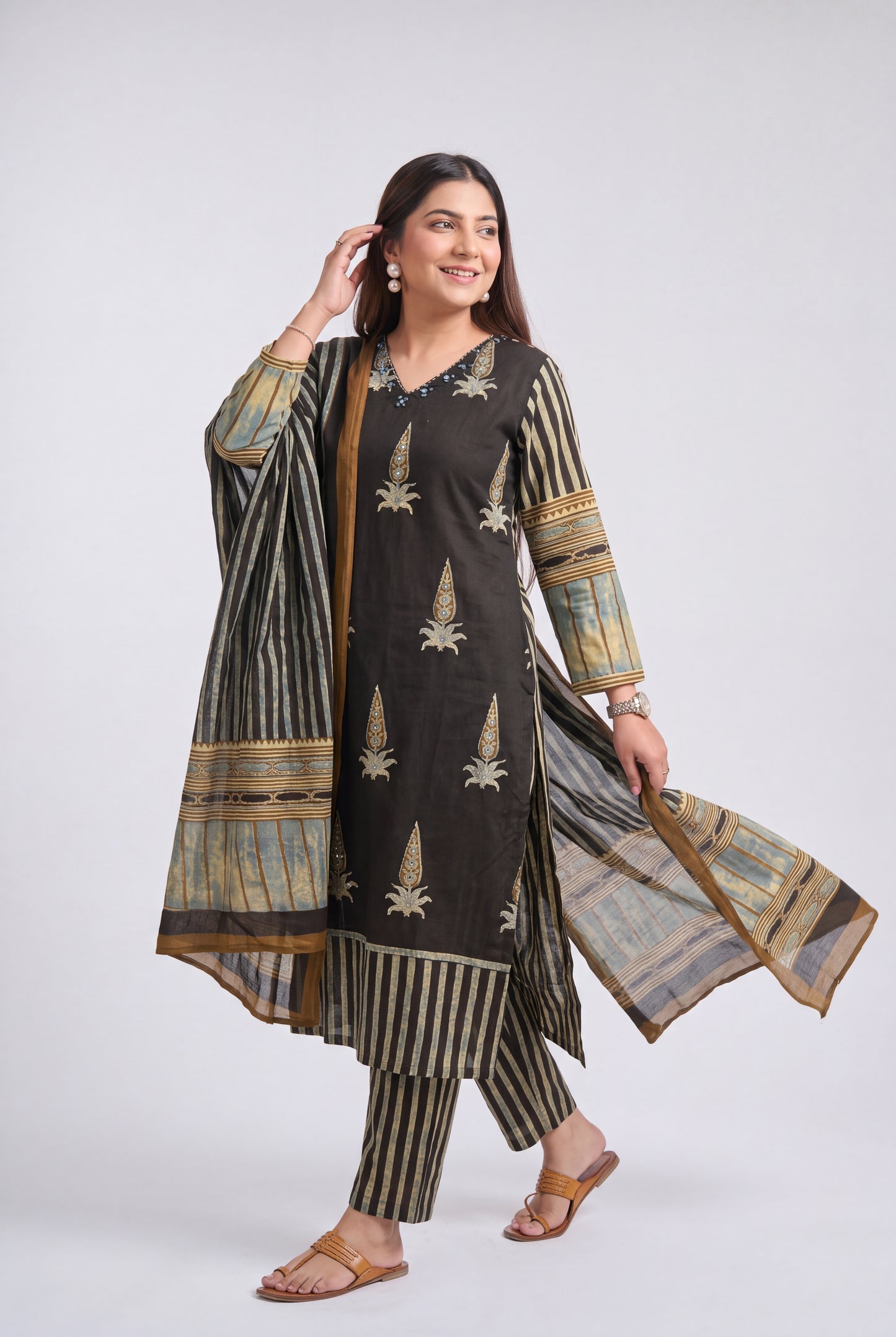 A woman modeling a traditional three-piece salwar kameez suit in dark brown and beige. The kurta features vertical beige stripes and embroidered motifs, paired with matching striped trousers and a sheer dupatta with beige and gold patterns, set against a plain light gray studio background.