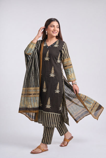 A woman modeling a traditional three-piece salwar kameez suit in dark brown and beige. The kurta features vertical beige stripes and embroidered motifs, paired with matching striped trousers and a sheer dupatta with beige and gold patterns, set against a plain light gray studio background.