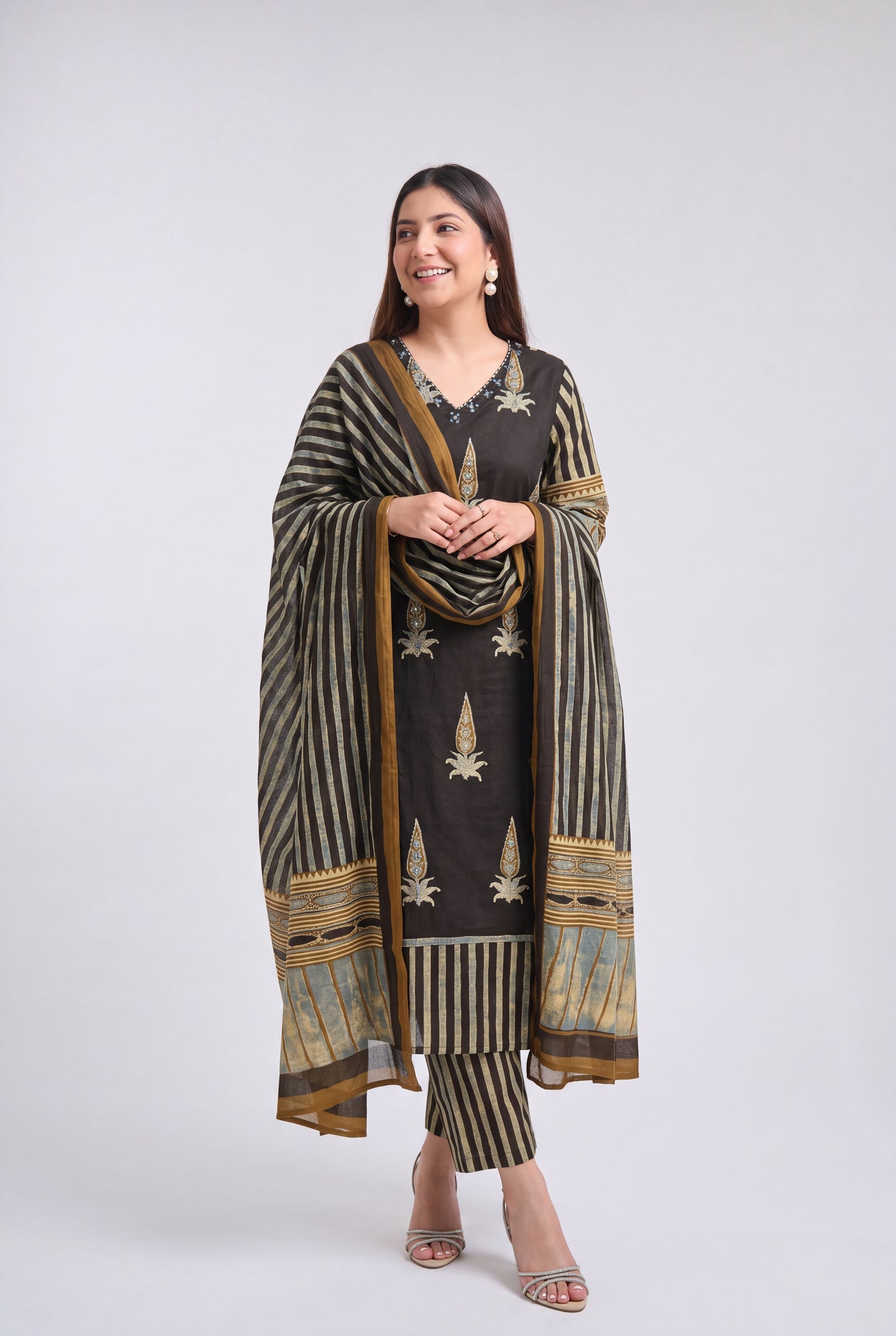 A woman modeling a traditional Indian salwar kameez suit set in dark brown and beige with gold and blue accents, featuring a striped dupatta and matching pants, against a light gray studio background.