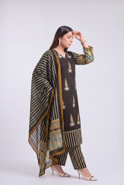 A woman modeling a three-piece ethnic suit set featuring a dark brown kurta with gold leaf motifs, matching striped pants, and a striped dupatta with gold borders, set against a plain light gray background.