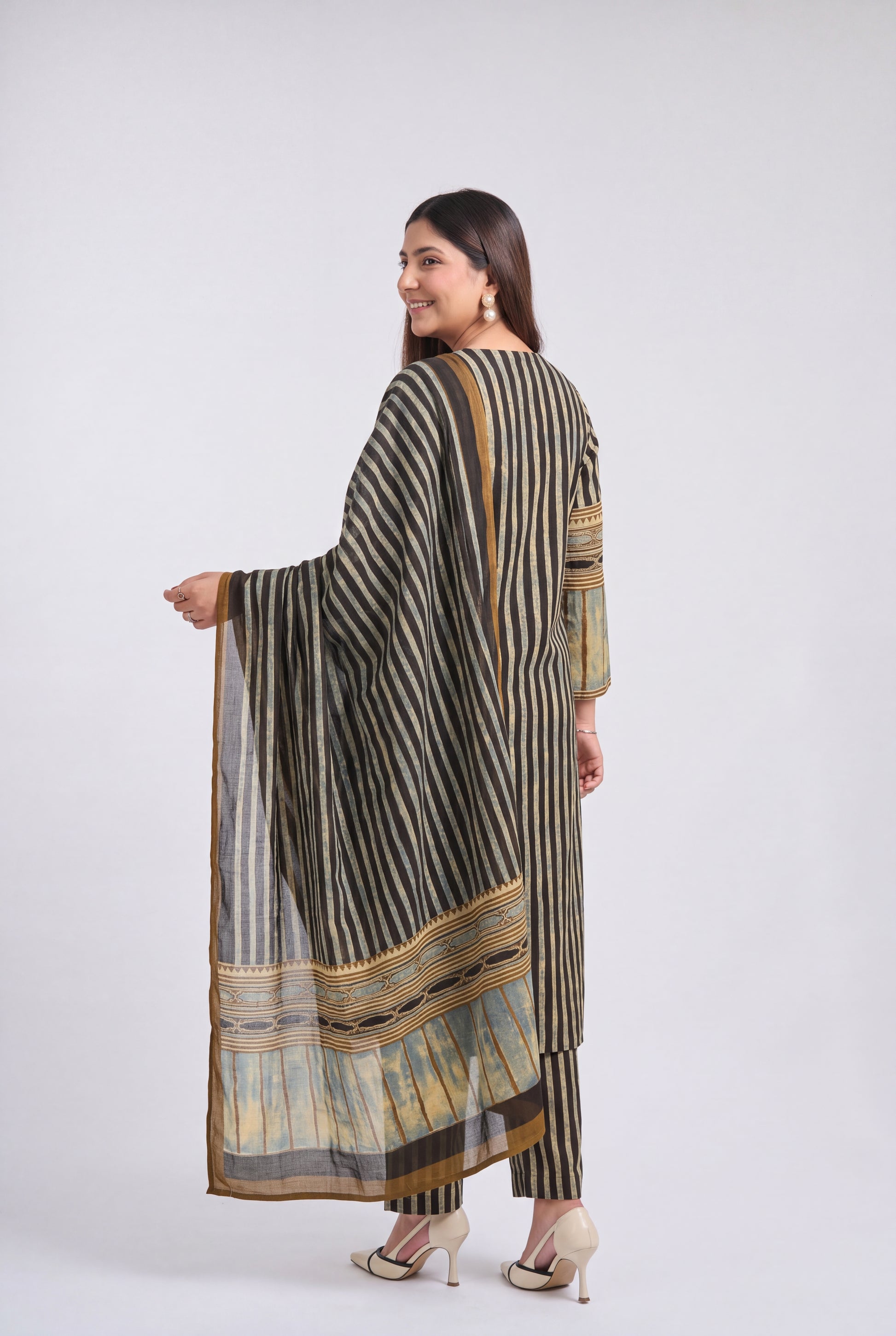 A woman modeling a three-piece traditional outfit with vertical stripes in black, beige, and brown, featuring a matching dupatta with a detailed border, photographed against a light gray studio background.