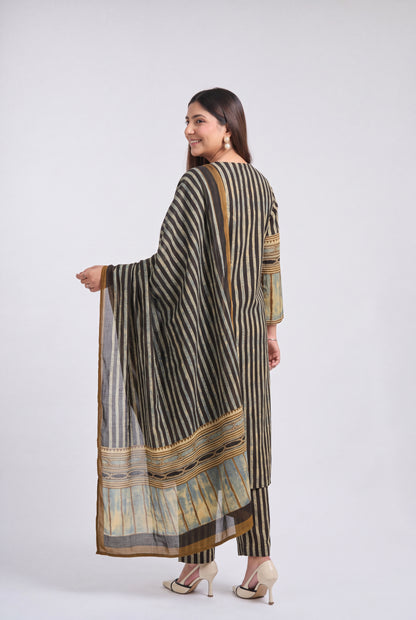 A woman modeling a three-piece traditional outfit with vertical stripes in black, beige, and brown, featuring a matching dupatta with a detailed border, photographed against a light gray studio background.