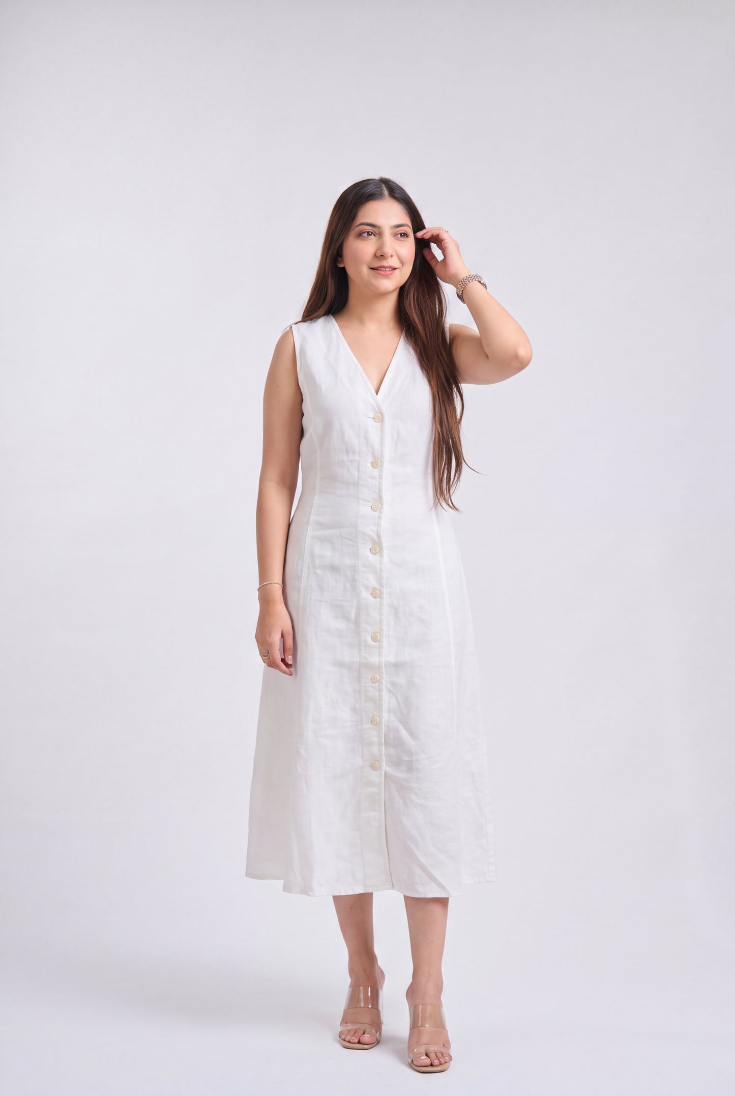 A woman modeling a sleeveless white linen midi dress with a V-neckline and a row of cream buttons down the front, standing against a light gray studio background.