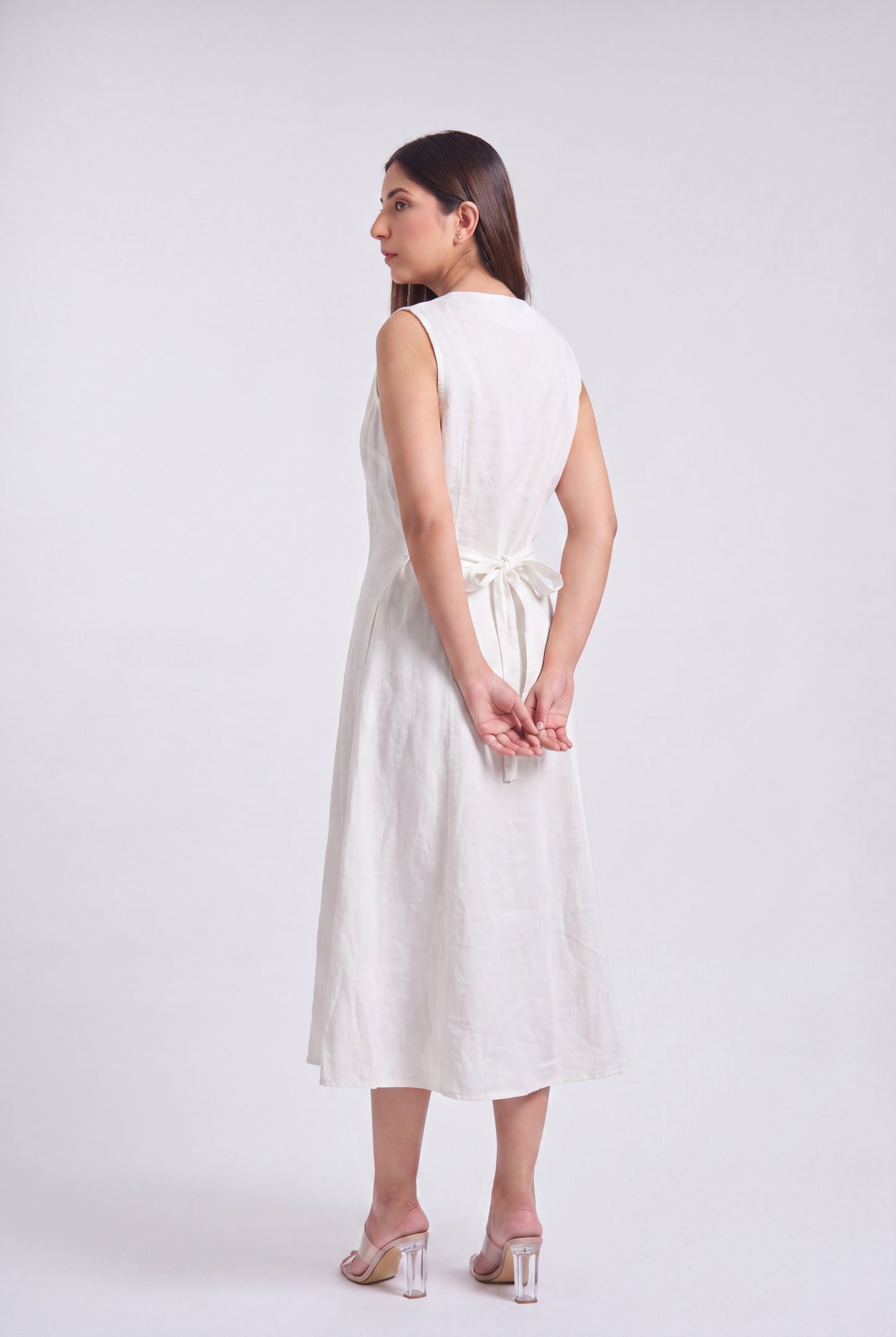 A woman modeling a sleeveless white linen midi dress from the back, featuring a tie waist detail, against a light gray studio background.