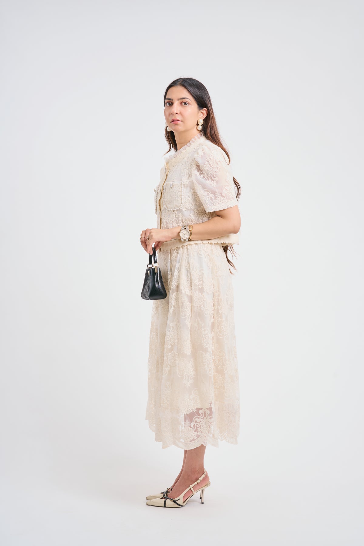 A woman modeling a cream lace midi dress with short puff sleeves and a tie waist, styled with a small black handbag and metallic heels against a plain white studio background.
