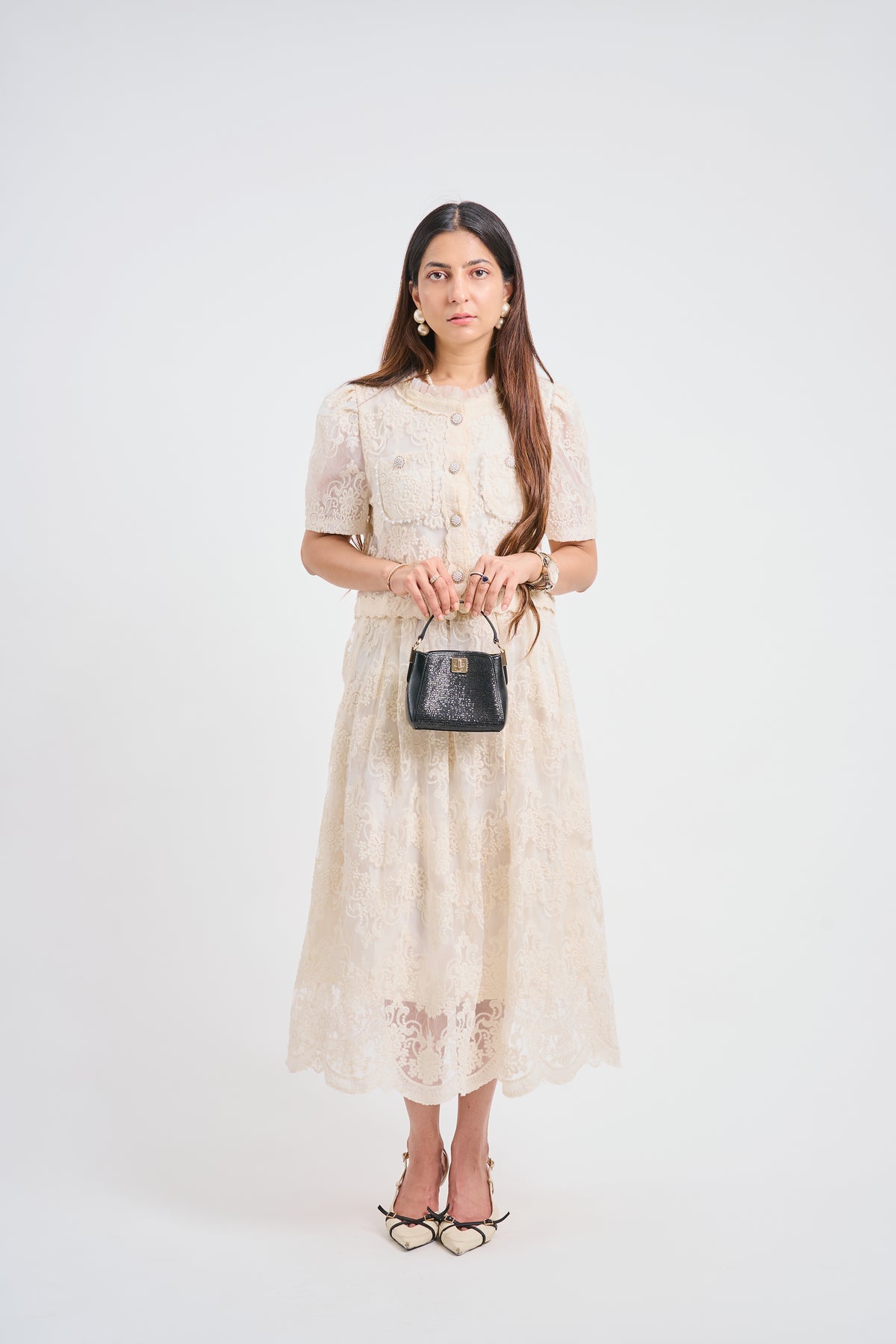 A woman stands against a plain light gray studio background wearing a cream lace midi dress with short sleeves, a round neckline, and front buttons. She is holding a small black handbag and wearing black strappy heels.