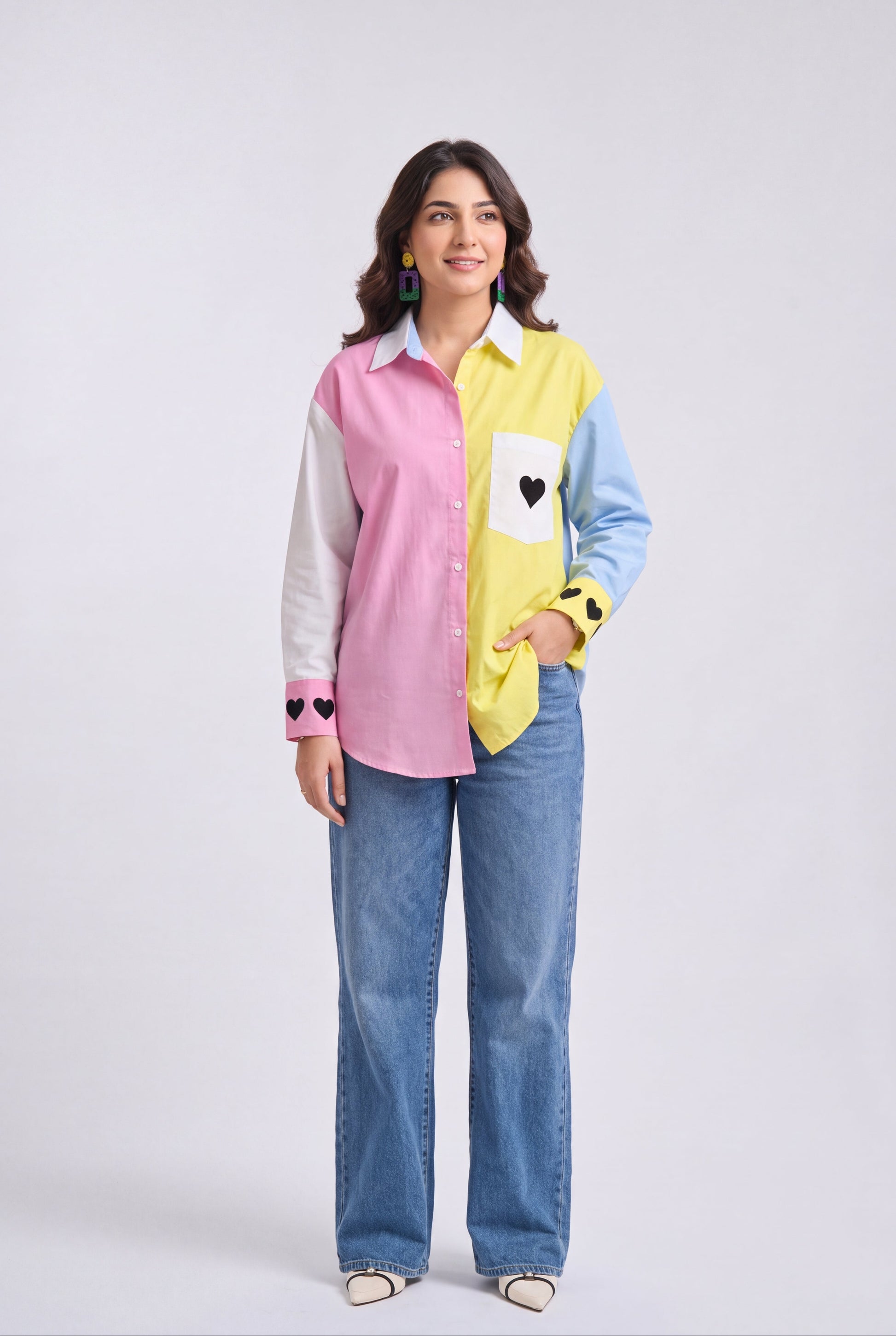 A woman modeling a color-blocked long-sleeve button-up shirt with pink, yellow, light blue, and white panels, paired with wide-leg blue jeans, against a light gray studio background.