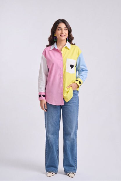 A woman modeling a color-blocked long-sleeve button-up shirt with pink, yellow, light blue, and white panels, paired with wide-leg blue jeans, against a light gray studio background.