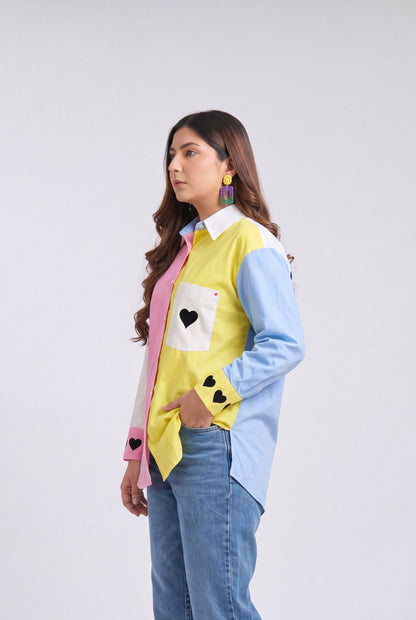 A woman modeling a colorful patchwork button-up shirt with blue, yellow, and pink panels, paired with wide-leg blue jeans and white heels, against a light gray studio background.