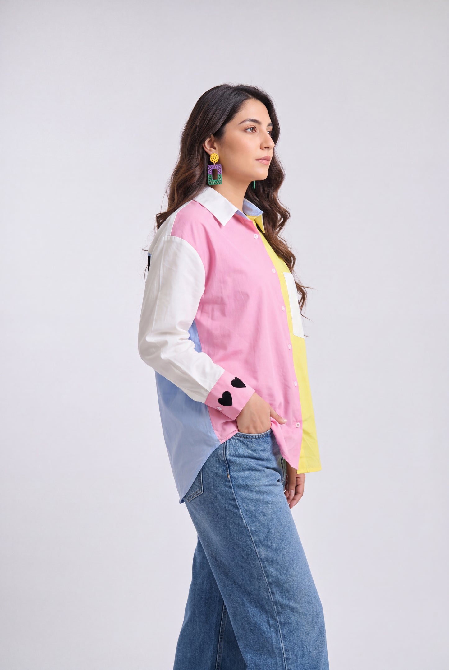 A woman modeling a color-blocked long-sleeve button-up shirt with pink, white, light blue, and yellow panels, paired with blue jeans, against a light gray studio background.