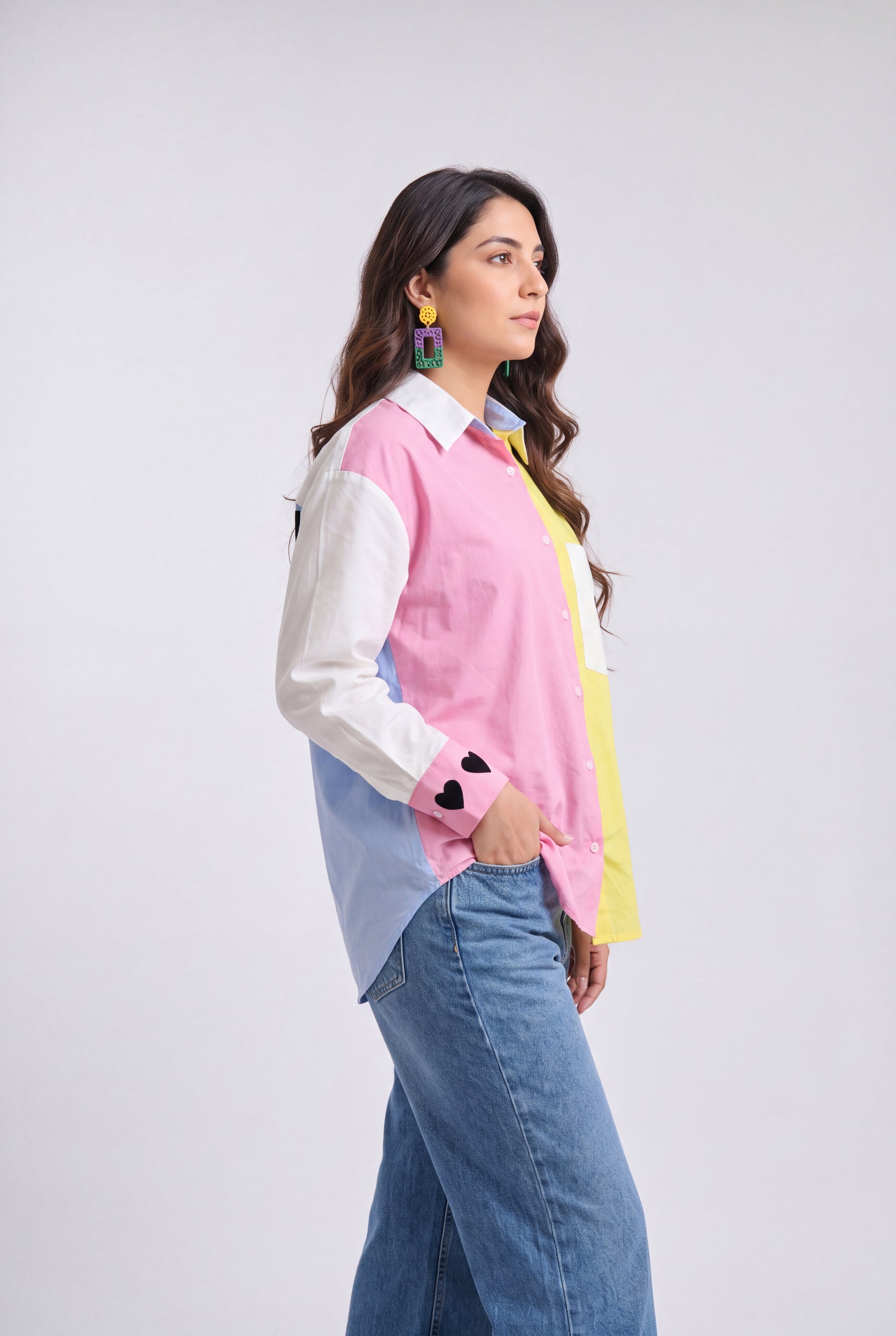A woman modeling a color-blocked long-sleeve button-up shirt with pink, white, light blue, and yellow panels, paired with blue jeans, against a light gray studio background.