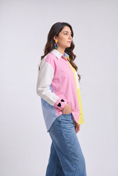 A woman modeling a color-blocked long-sleeve button-up shirt with pink, white, light blue, and yellow panels, paired with blue jeans, against a light gray studio background.