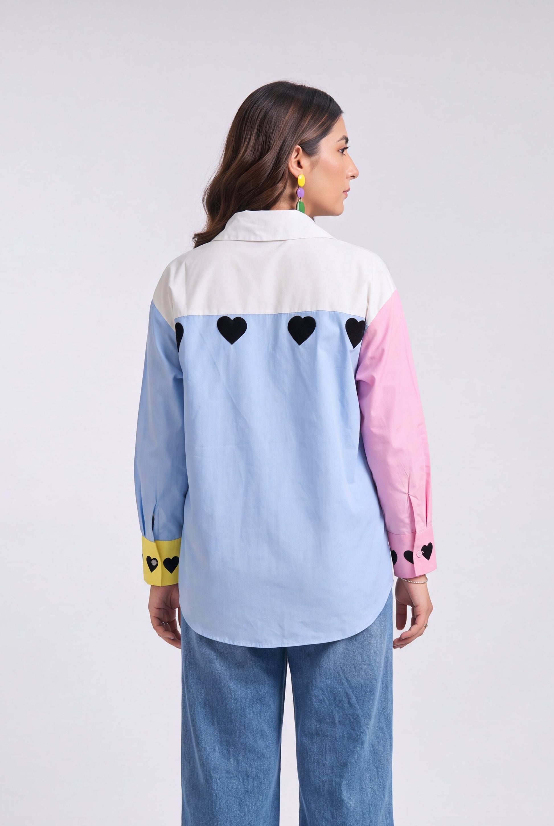 A model shown from the back wearing a light blue, pink, and white color-blocked shirt with black heart details, paired with wide-leg blue jeans, against a light gray studio background.