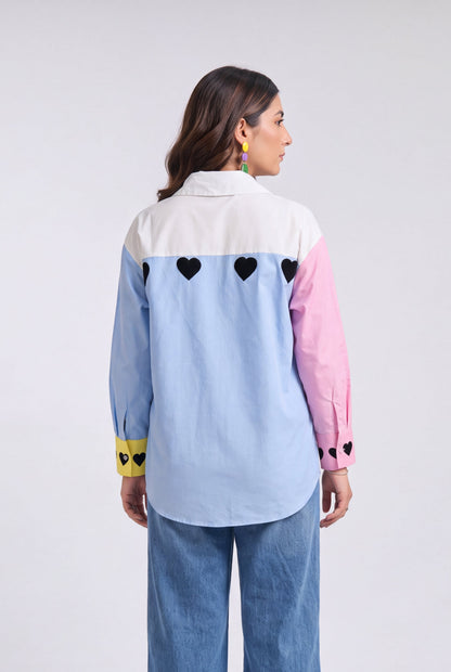 A model shown from the back wearing a light blue, pink, and white color-blocked shirt with black heart details, paired with wide-leg blue jeans, against a light gray studio background.