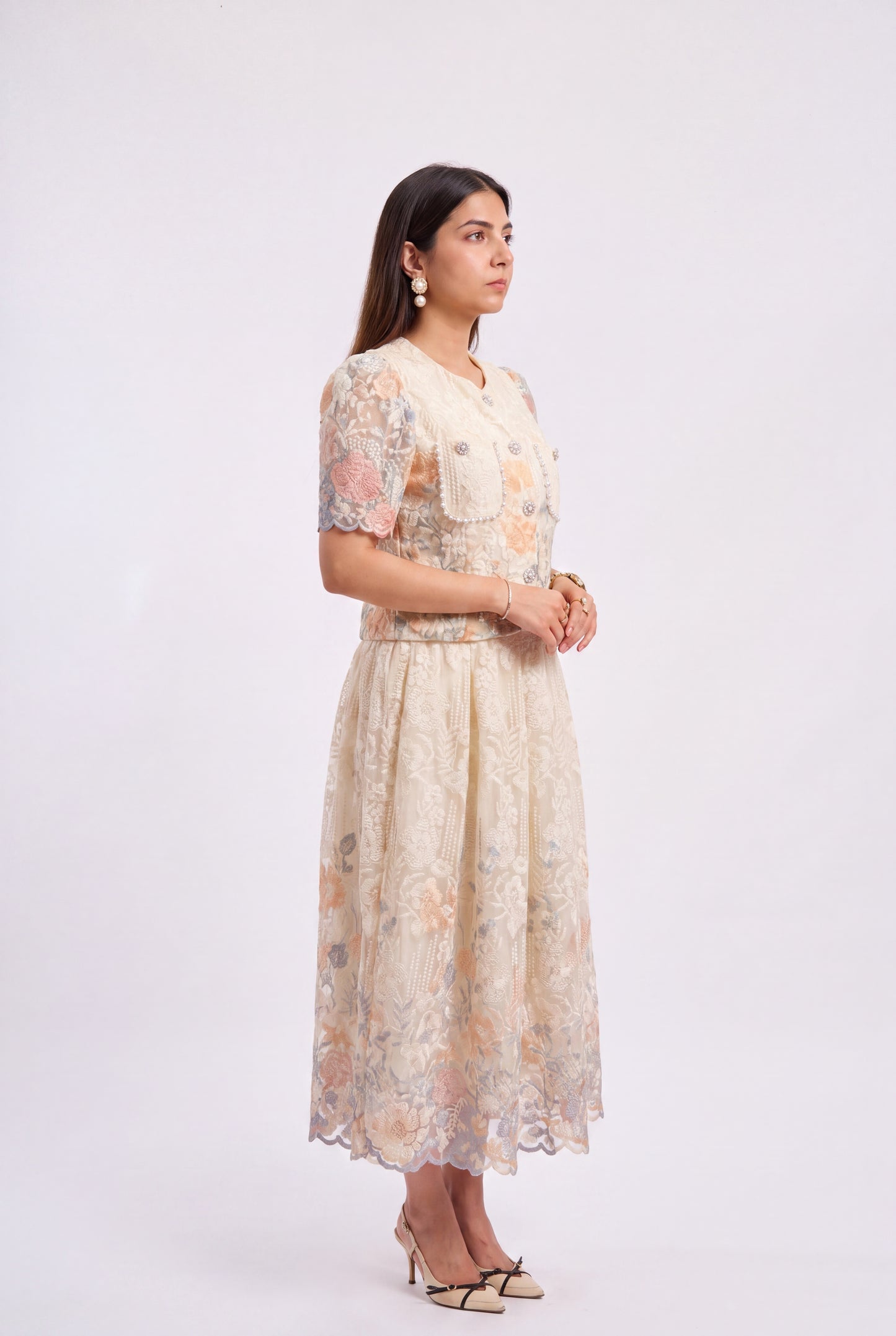 A woman modeling a cream-colored lace midi dress with peach and gray floral patterns, standing against a plain white studio background.