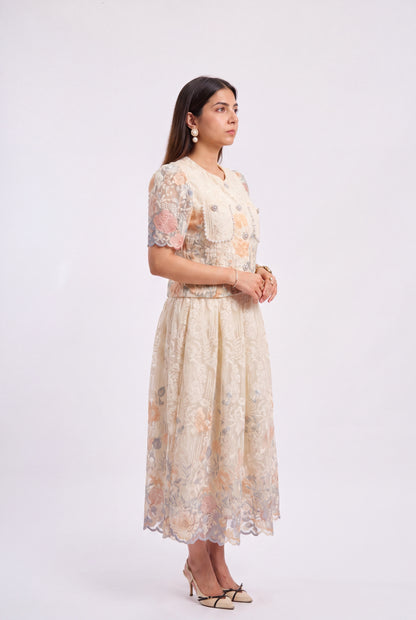 A woman modeling a cream-colored lace midi dress with peach and gray floral patterns, standing against a plain white studio background.