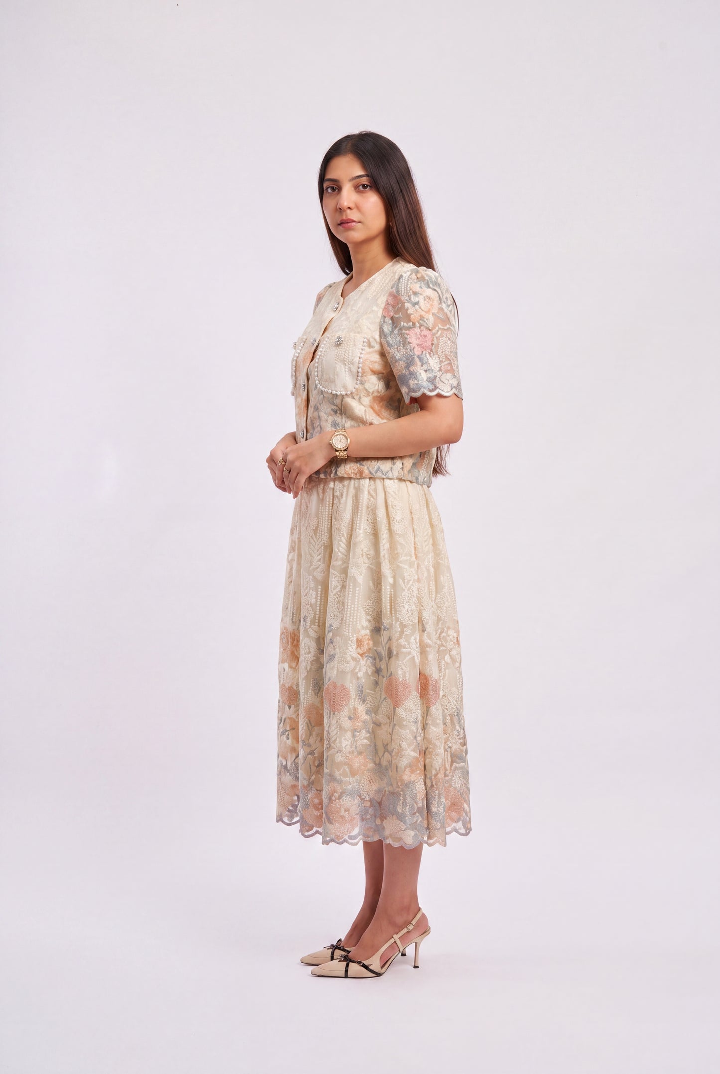 A woman modeling a short-sleeved midi dress with a beige base and a soft floral pattern in muted pink, peach, and blue, standing against a plain light gray studio background.