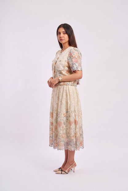A woman modeling a short-sleeved midi dress with a beige base and a soft floral pattern in muted pink, peach, and blue, standing against a plain light gray studio background.