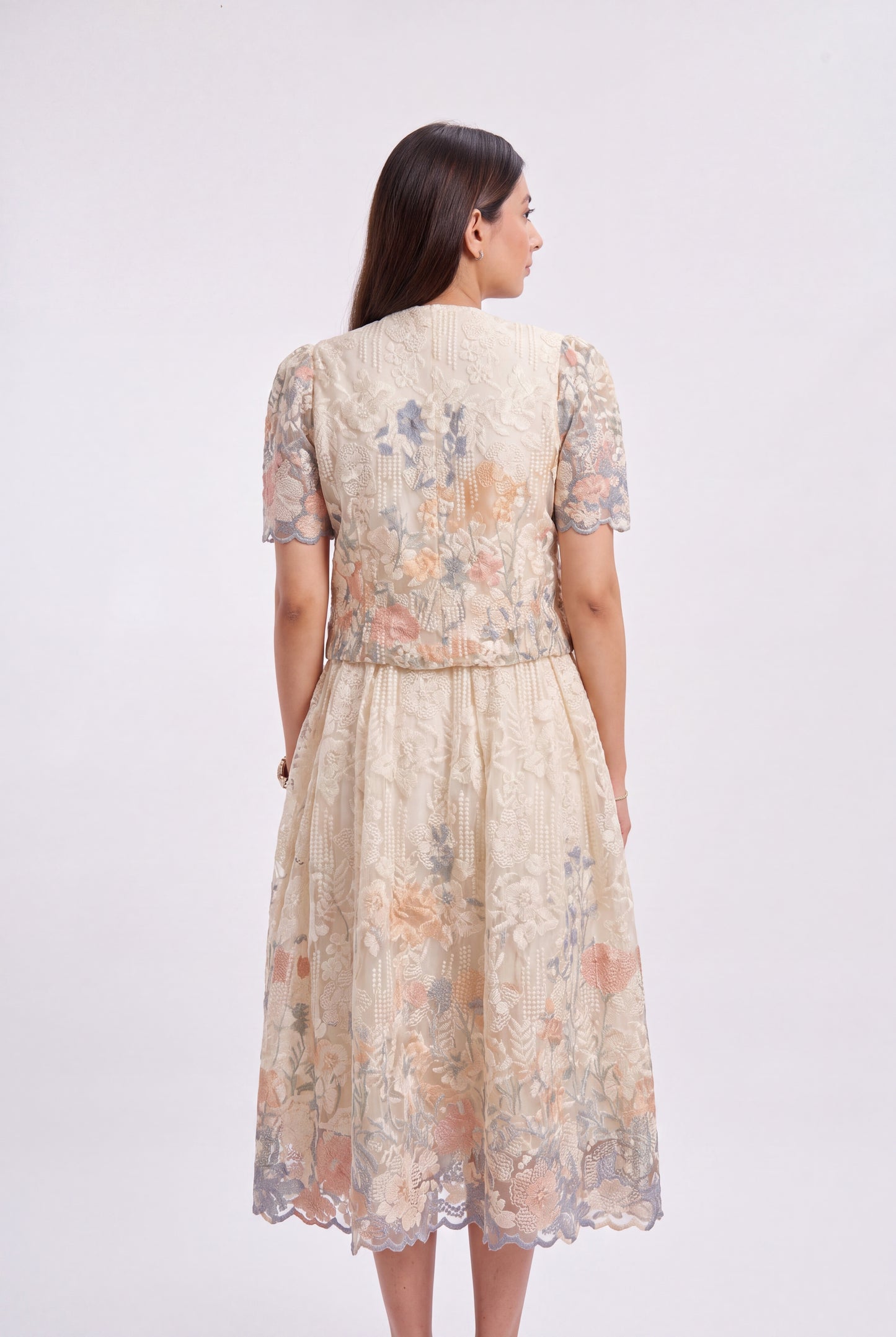 A woman viewed from the back wearing a short-sleeved midi dress with a textured floral lace pattern in beige, peach, and muted blue-gray tones, set against a plain light gray studio background.