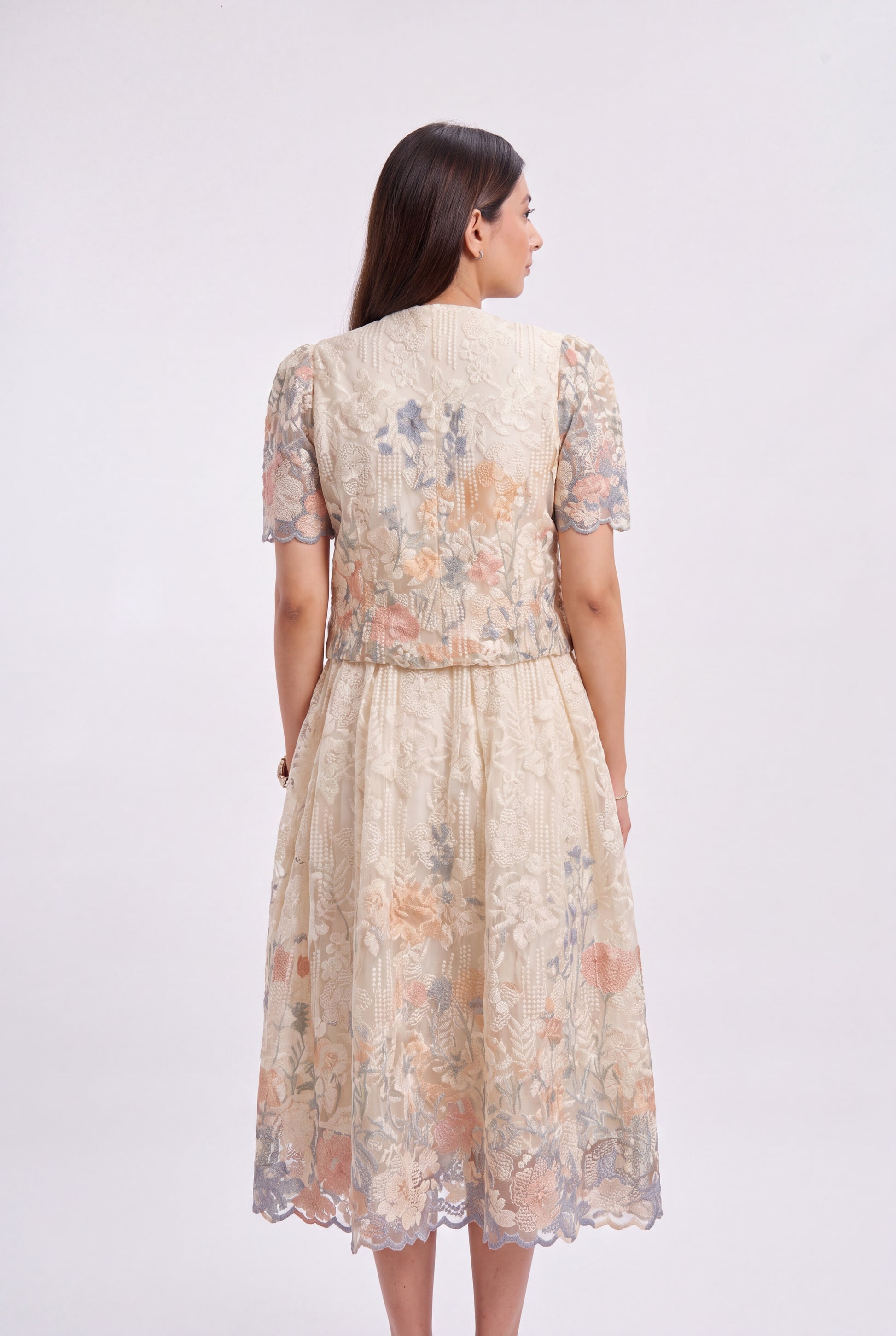 A woman viewed from the back wearing a short-sleeved midi dress with a textured floral lace pattern in beige, peach, and muted blue-gray tones, set against a plain light gray studio background.